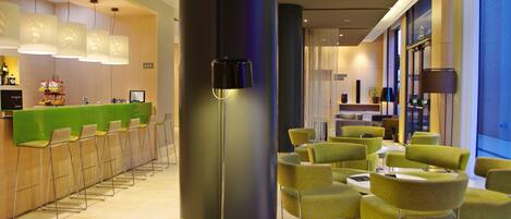 A modern hotel lobby with a bar area, green upholstered chairs, and a large floor lamp.