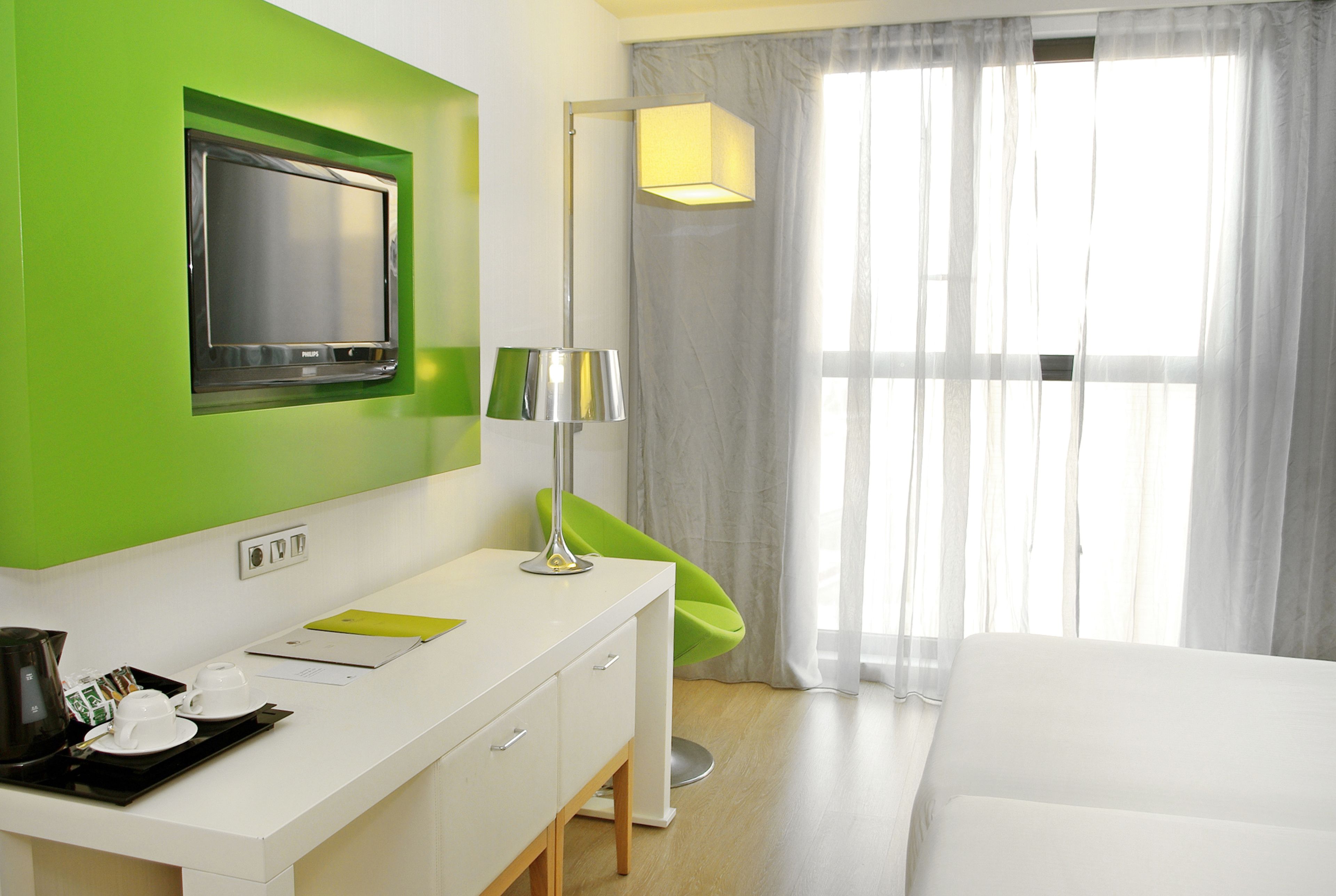 A hotel room with a green accent wall, a flat-screen TV, a desk with a lamp, a bed, and a window with sheer curtains.