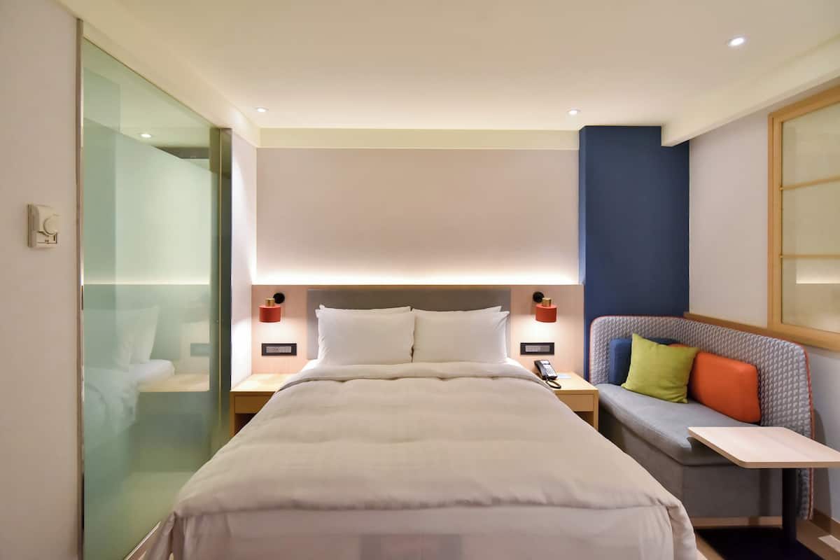 Een hotelkamer met een bed, nachtkastjes, een zithoek met kussens en een glazen scheidingswand.