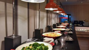A buffet line with various dishes, including a red bowl of broccoli and carrots, and a black speaker in the background.