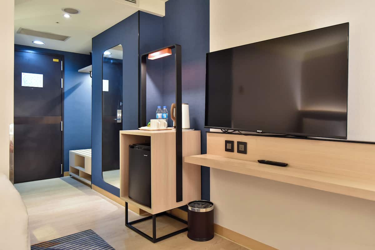 Een moderne hotelkamer met een groot flatscreen televisietoestel, een minibar, een prullenbak en een spiegel.