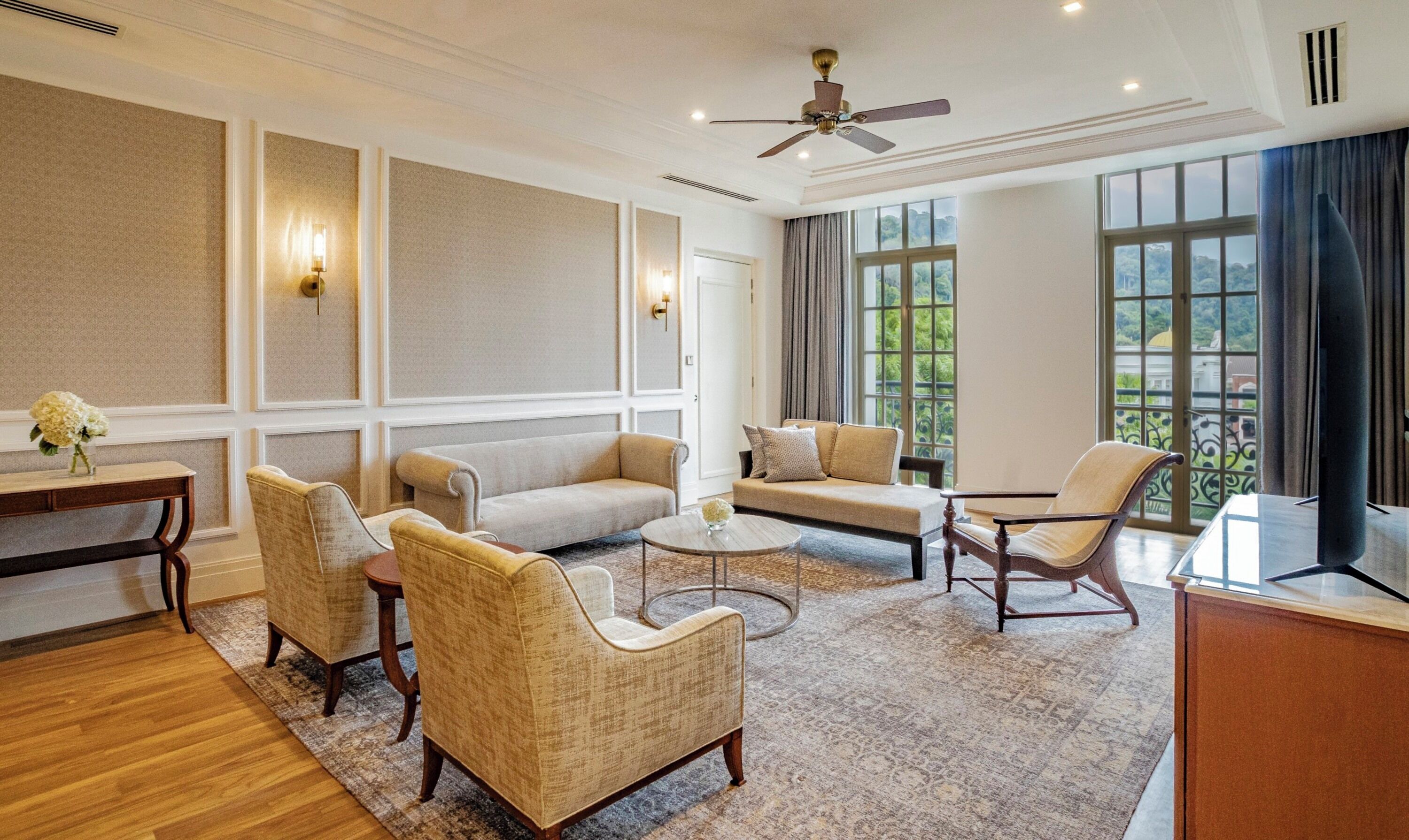 A spacious living room with a high ceiling, a ceiling fan, and large windows offering a view of greenery outside.
