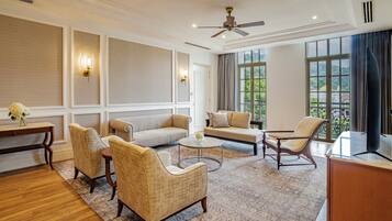 A spacious living room with a high ceiling, a ceiling fan, and large windows offering a view of greenery outside.