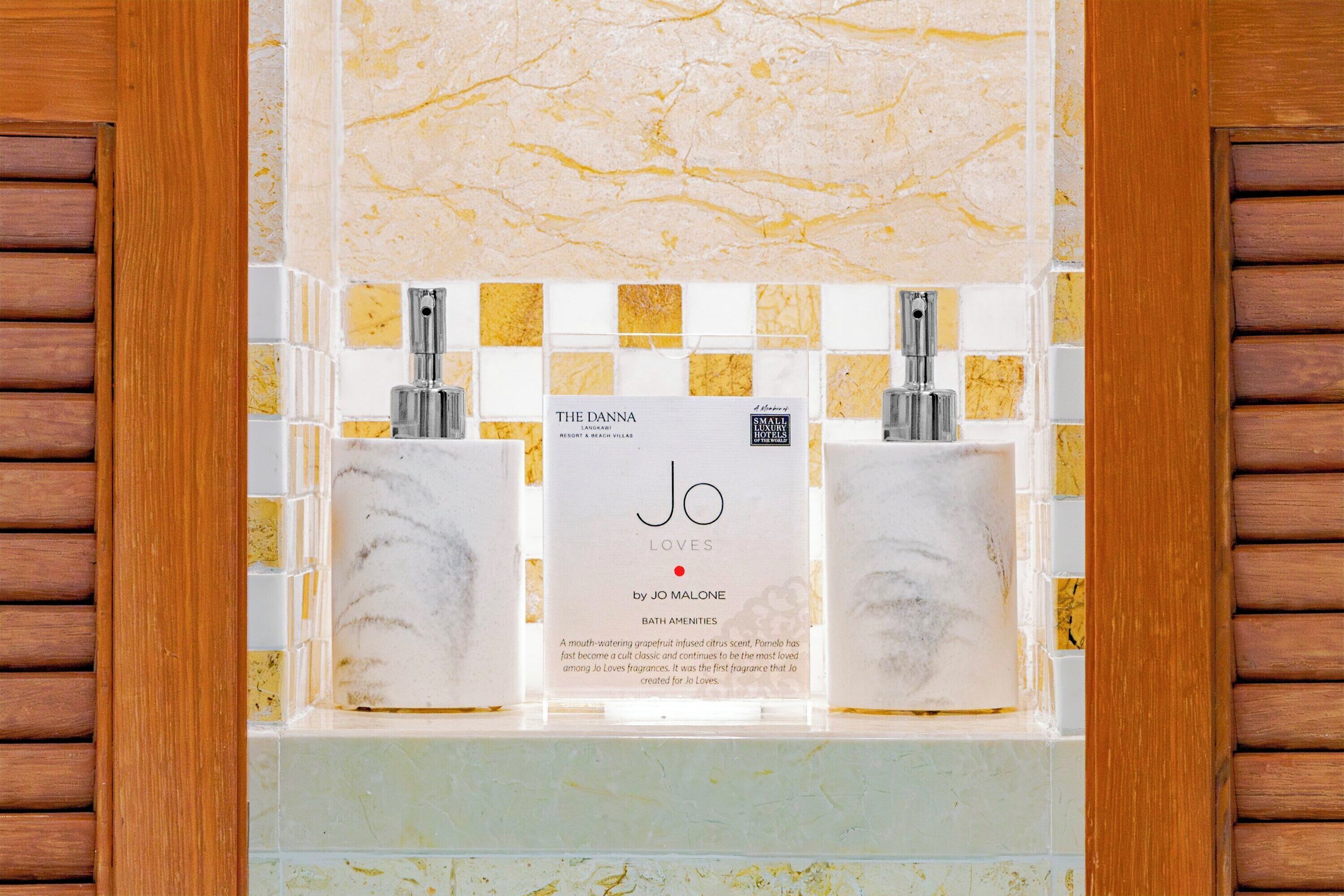 A bathroom with a marble sink, two glass soap dispensers, and a sign for 'The Danna' and 'Jo Loves by Jo Malone Bath Amenities'.