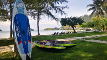 A beachside setting with a surfboard leaning against a tree, a kayak, and lounge chairs under umbrellas.