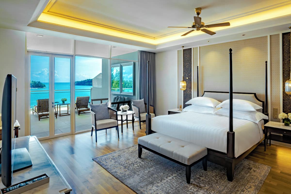 A spacious bedroom with a large bed, a ceiling fan, and a view of the sea.