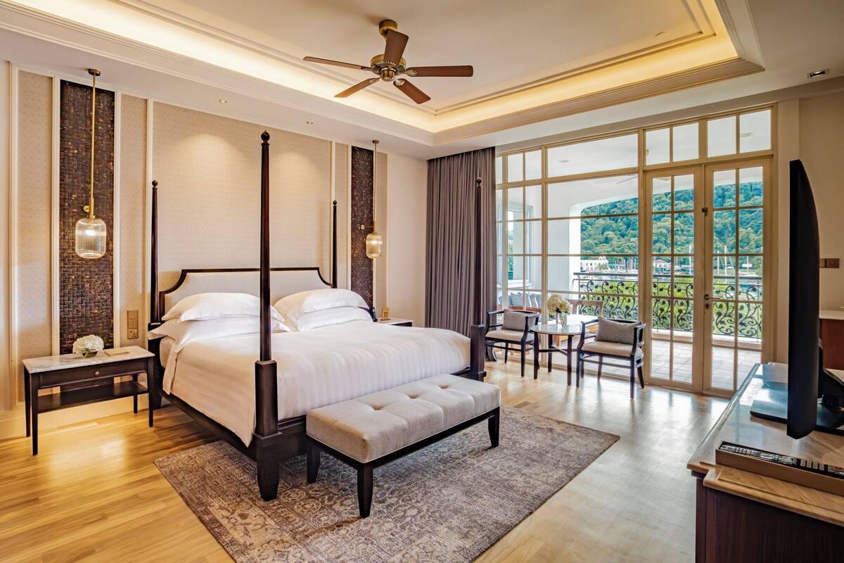 A spacious bedroom with a large bed, a ceiling fan, a view of the outdoors through large windows, and a small seating area with a table and chairs.