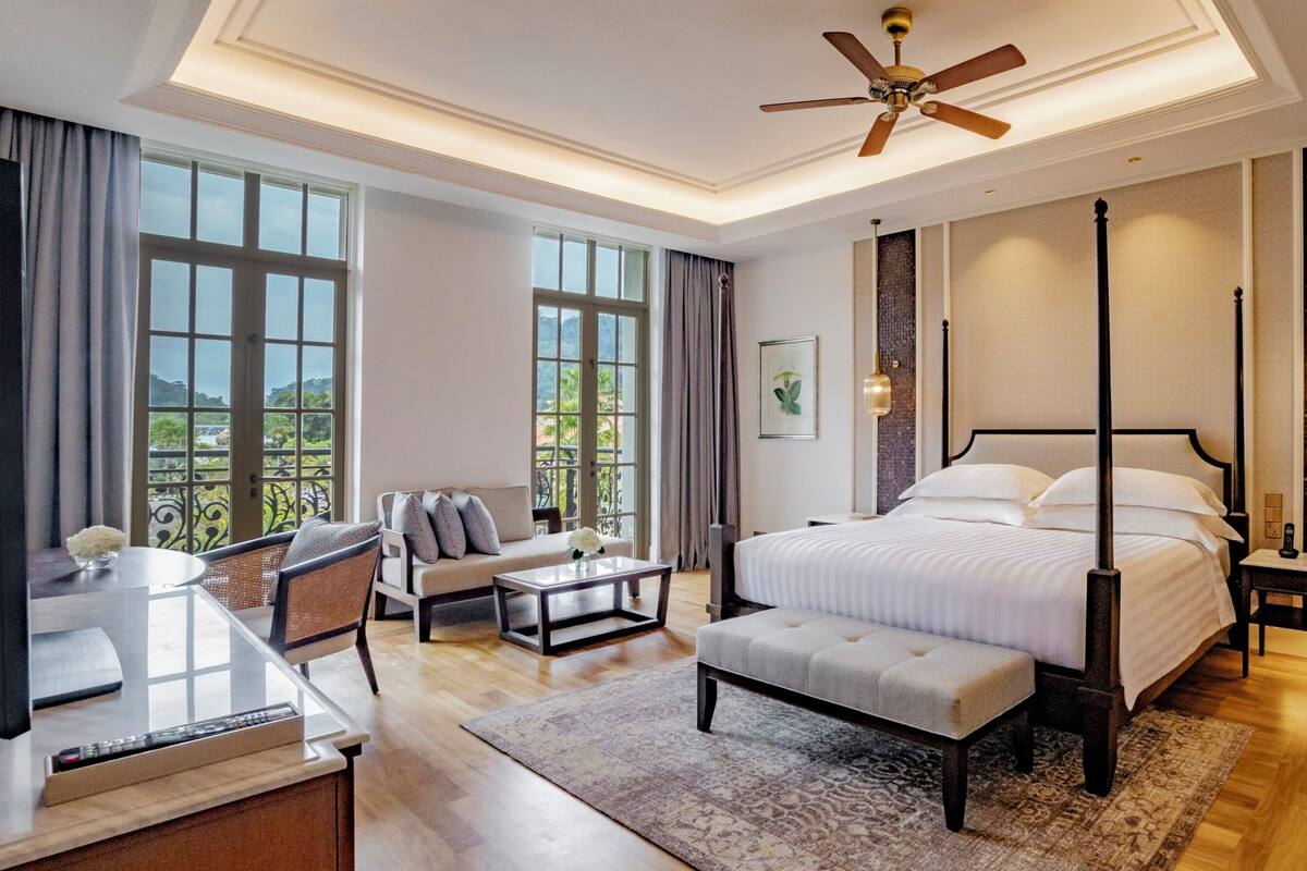 A modern hotel room with a large bed, a desk with a chair, a sofa, and a balcony with a view of greenery.