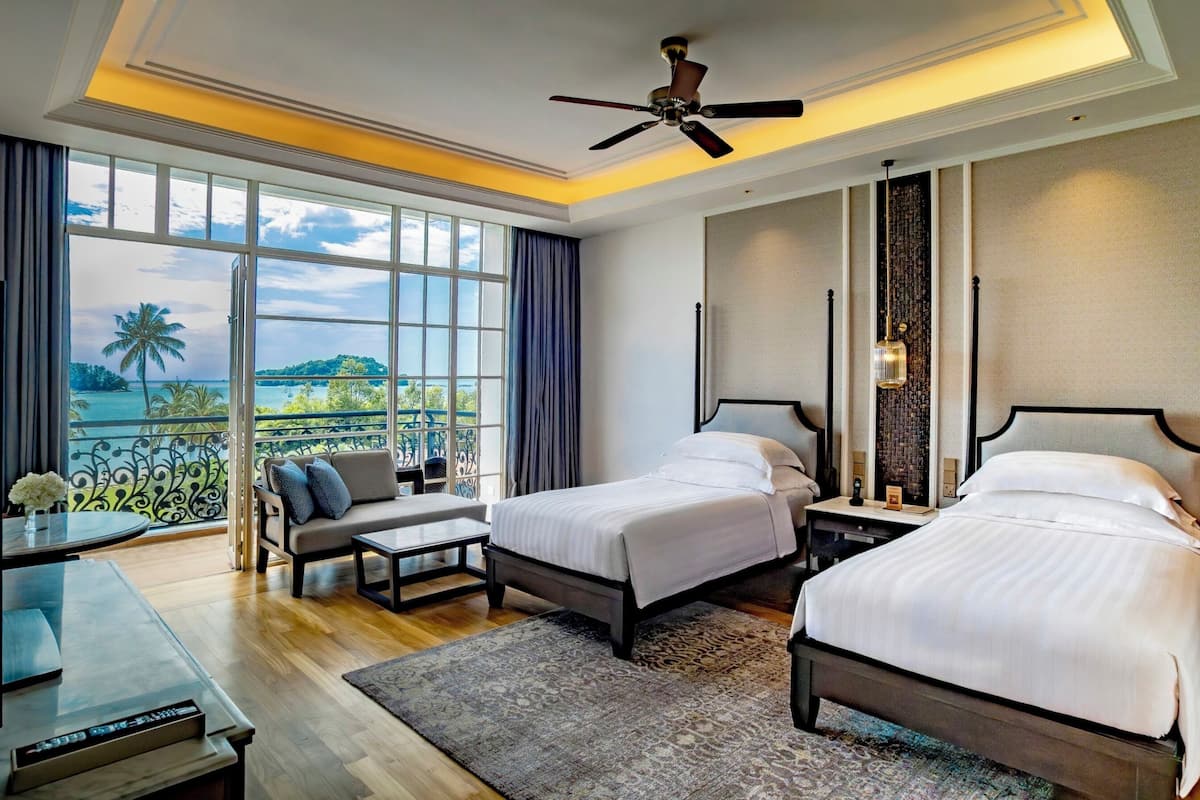 A hotel room with two beds, a desk, a chair, a ceiling fan, and a view of the ocean.