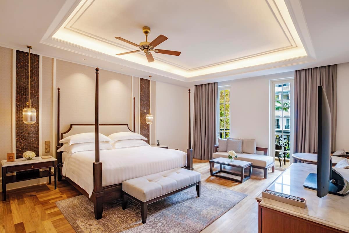 A spacious bedroom with a four-poster bed, a sitting area with a sofa and coffee table, a television, and a balcony with a view.
