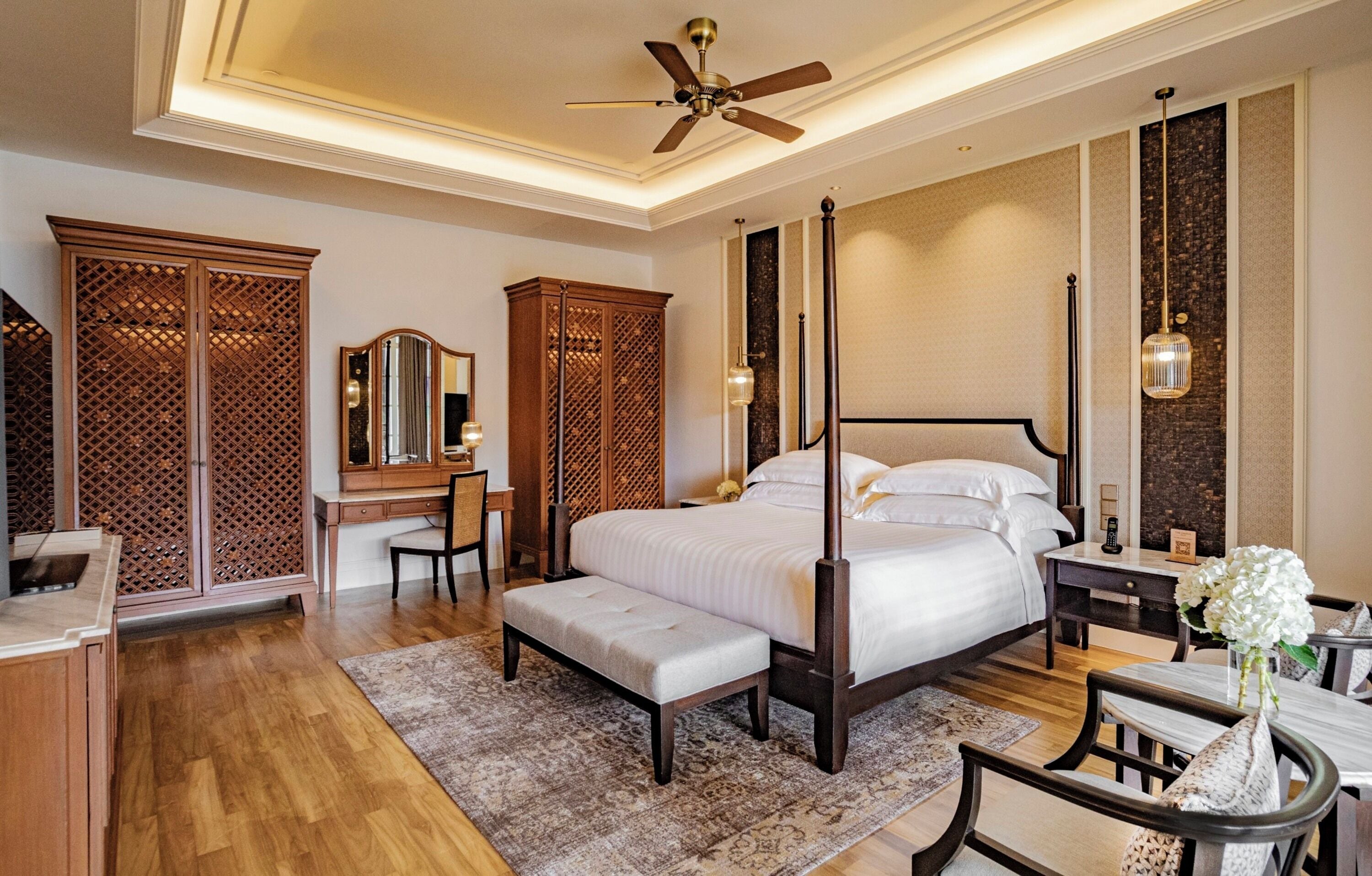 A spacious bedroom with a four-poster bed, a desk with a chair, a vanity with a mirror, and a wardrobe.