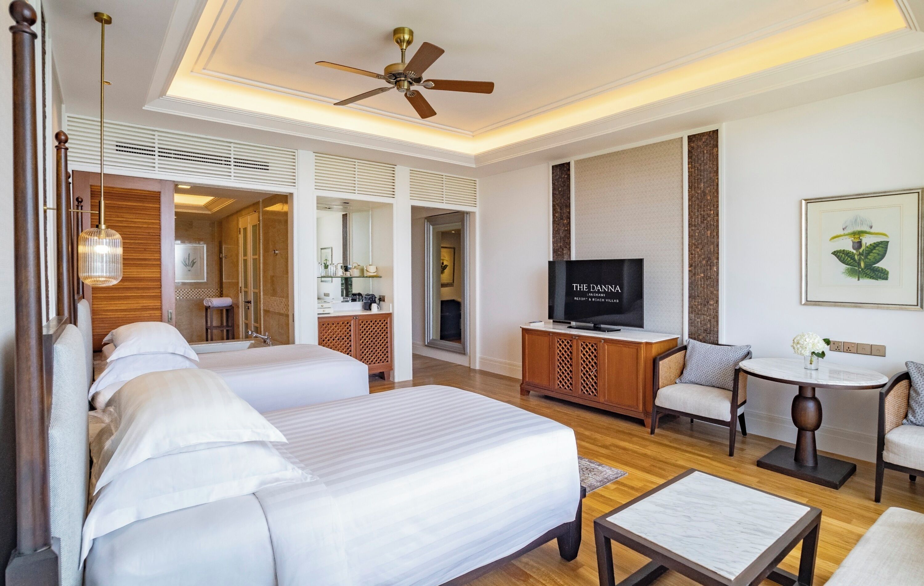 Habitación de hotel con una cama grande, televisor, zona de estar con sillas, una mesita y un ventilador de techo.