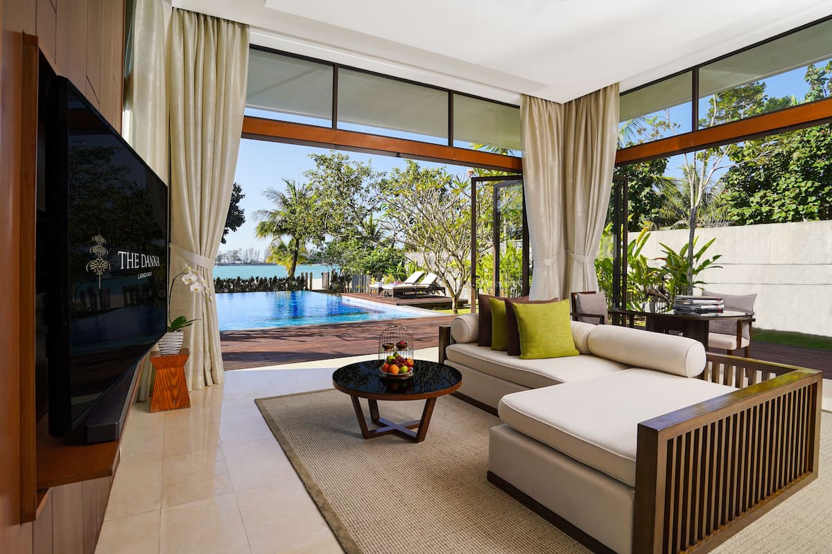 A modern living room with a large TV, a sofa, a coffee table with fruit, and a view of a swimming pool and greenery.