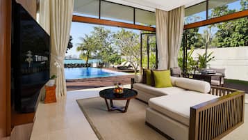 Una sala de estar moderna con un televisor grande, un sofá, una mesa de centro con fruta y vistas a una piscina y zonas verdes.