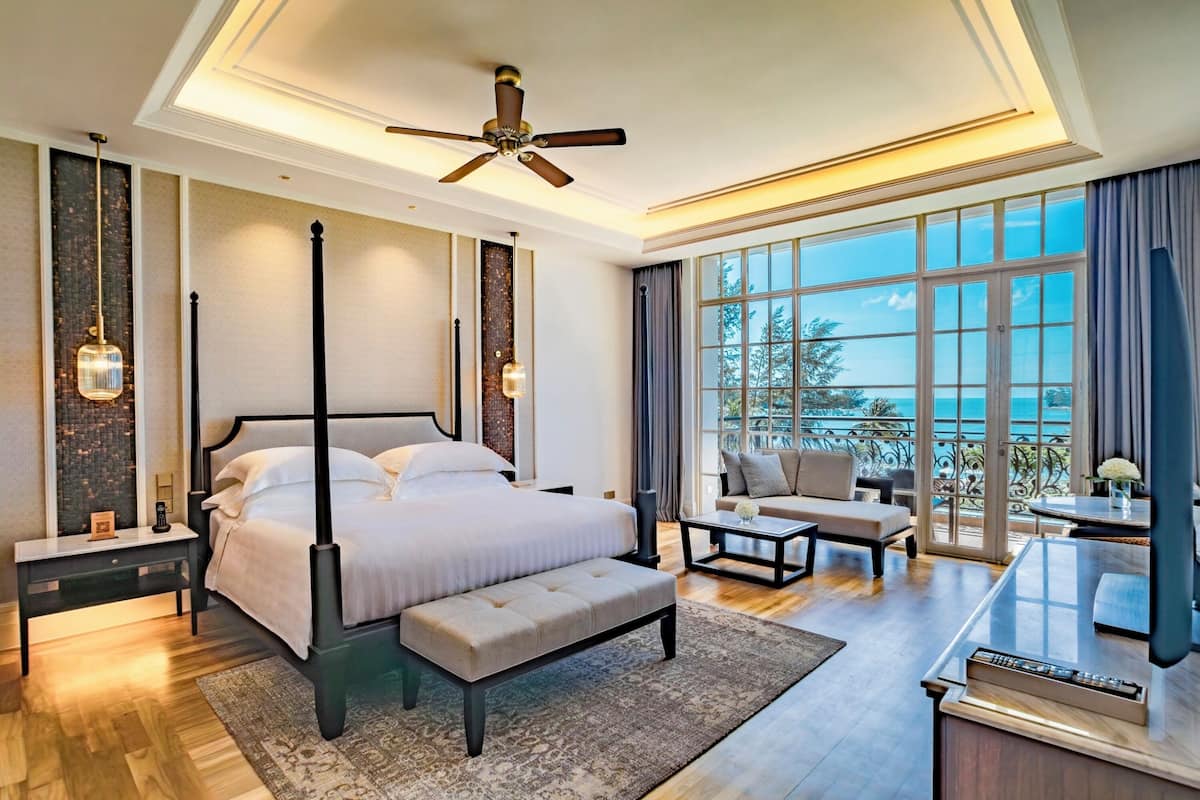 A spacious bedroom with a large bed, a ceiling fan, a view of the outdoors, and a seating area with a table and chairs.