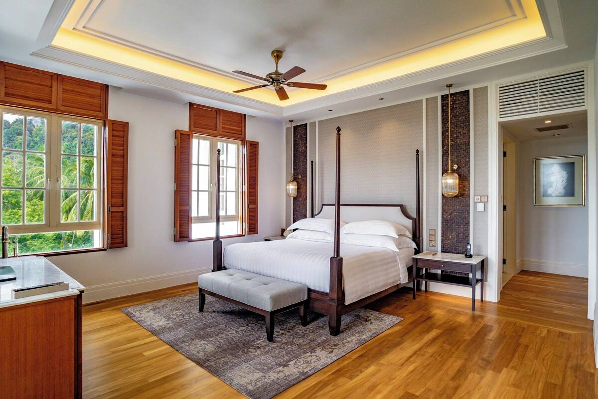 A bedroom with a four-poster bed, a wooden floor, a large window with shutters, and a ceiling fan.