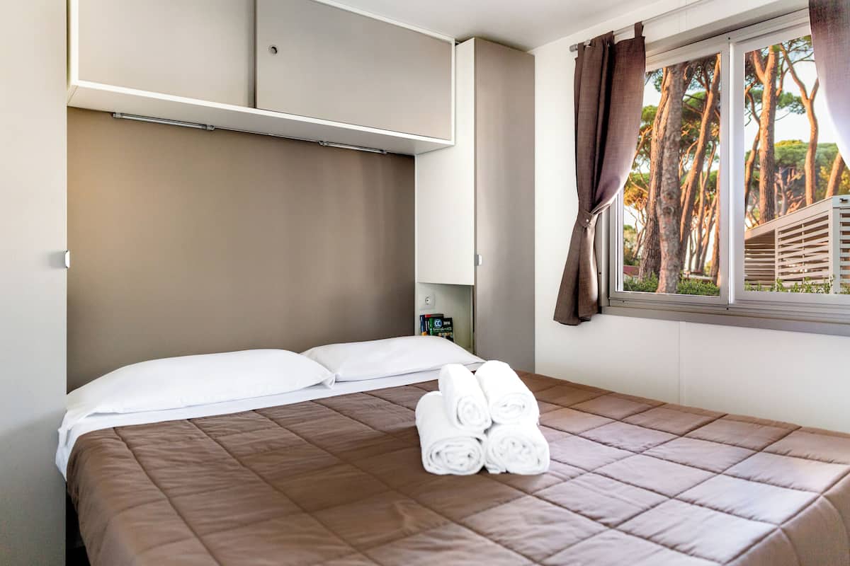 A neatly made bed with a brown quilt, a set of rolled towels, and a window with curtains.