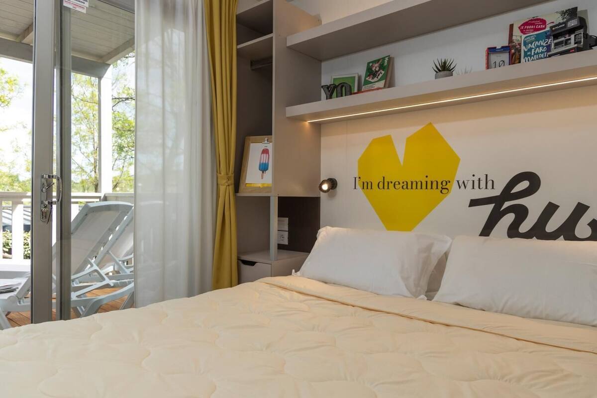 A bedroom with a bed, a window with curtains, a shelf with books and a framed picture, and a wall with a heart-shaped decoration and text.