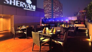 An outdoor dining area with tables and chairs, a sign for 'SHERVANI', and a building in the background. - Shervani Hotel Nehru Place (New Delhi)