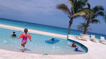 A pool area with clear blue water, a sandy beach, and palm trees.