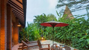 Un patio al aire libre con mobiliario de madera y un paraguas rojo, rodeado de exuberante follaje y una estructura cónica tradicional al fondo.