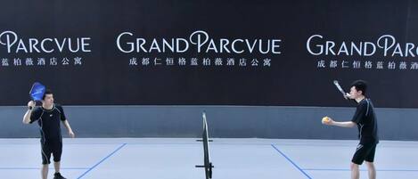A tennis court with two players practicing. The court has blue lines marking the boundaries. There is a black net in the center. The background features a wall with the text "Grand Parcvue" and "Blue Bamboo Hotel".