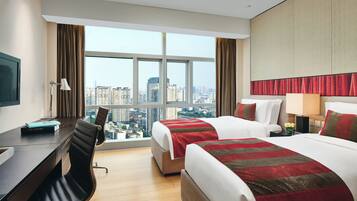 A hotel room with a desk, two beds, a chair, and a large window offering a city view.