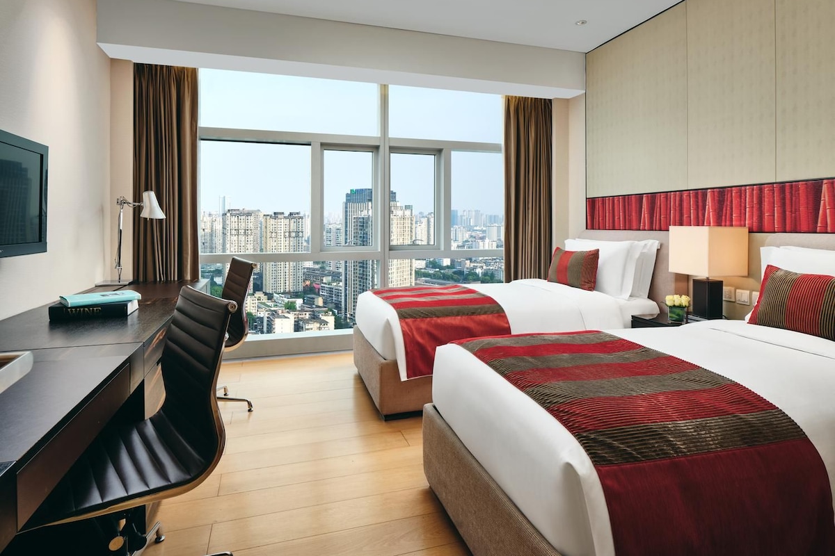 Habitación de hotel con escritorio, dos camas, televisión y vista a la ciudad.