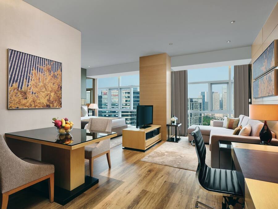 Habitación de hotel moderna con zona de comedor, televisión, sofá y ventana grande con vistas a la ciudad.