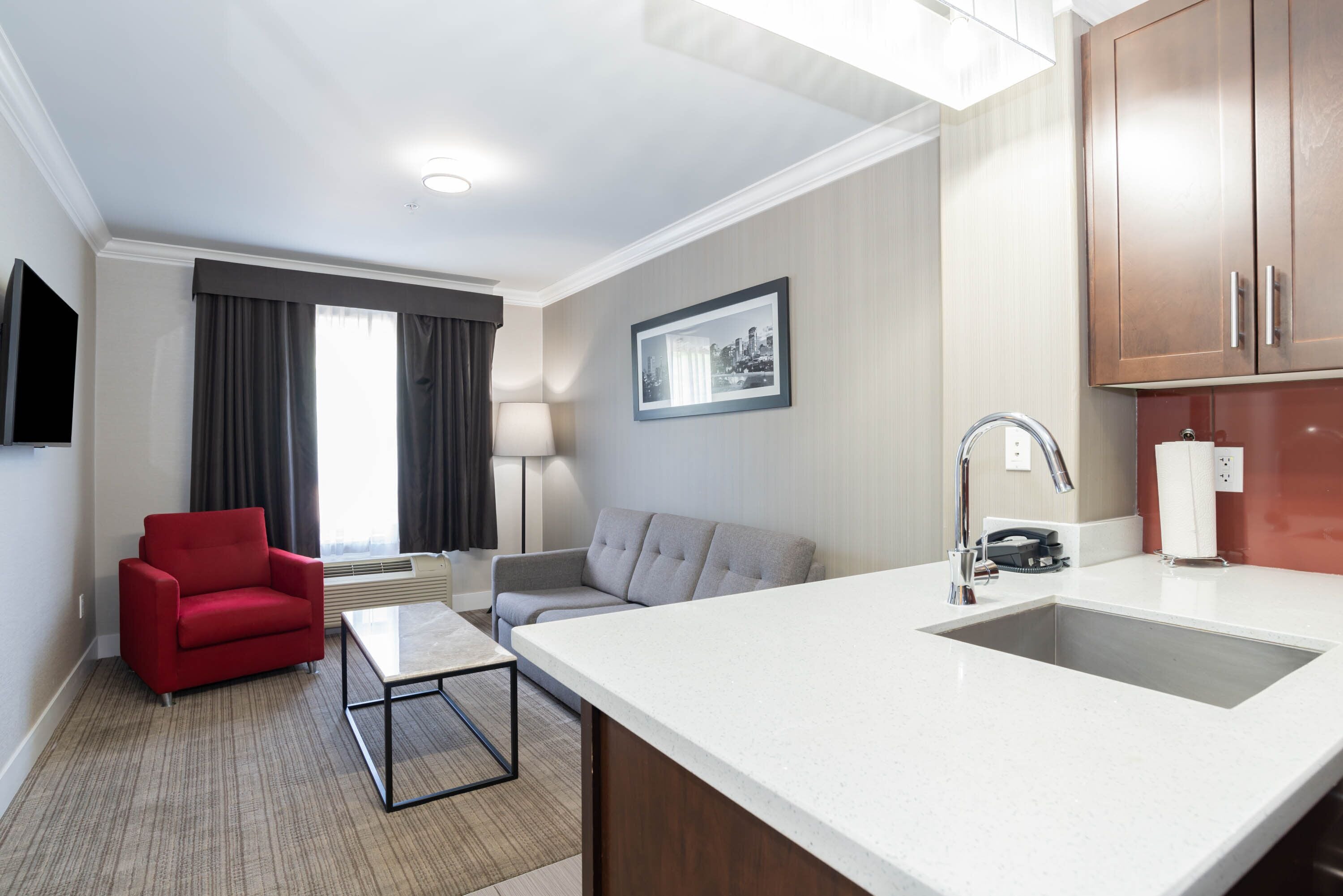 Quarto de hotel moderno com cozinha compacta, uma poltrona vermelha, sofá cinza, mesa de centro e uma grande janela com cortinas.