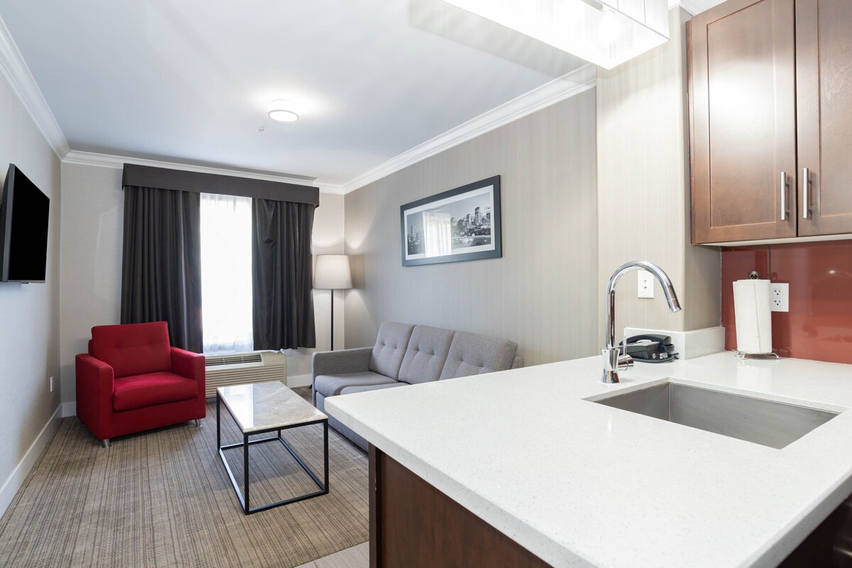 Una habitación de hotel moderna con kitchenette, un sillón rojo, un sofá gris, una mesa de centro y un ventanal con cortinas.
