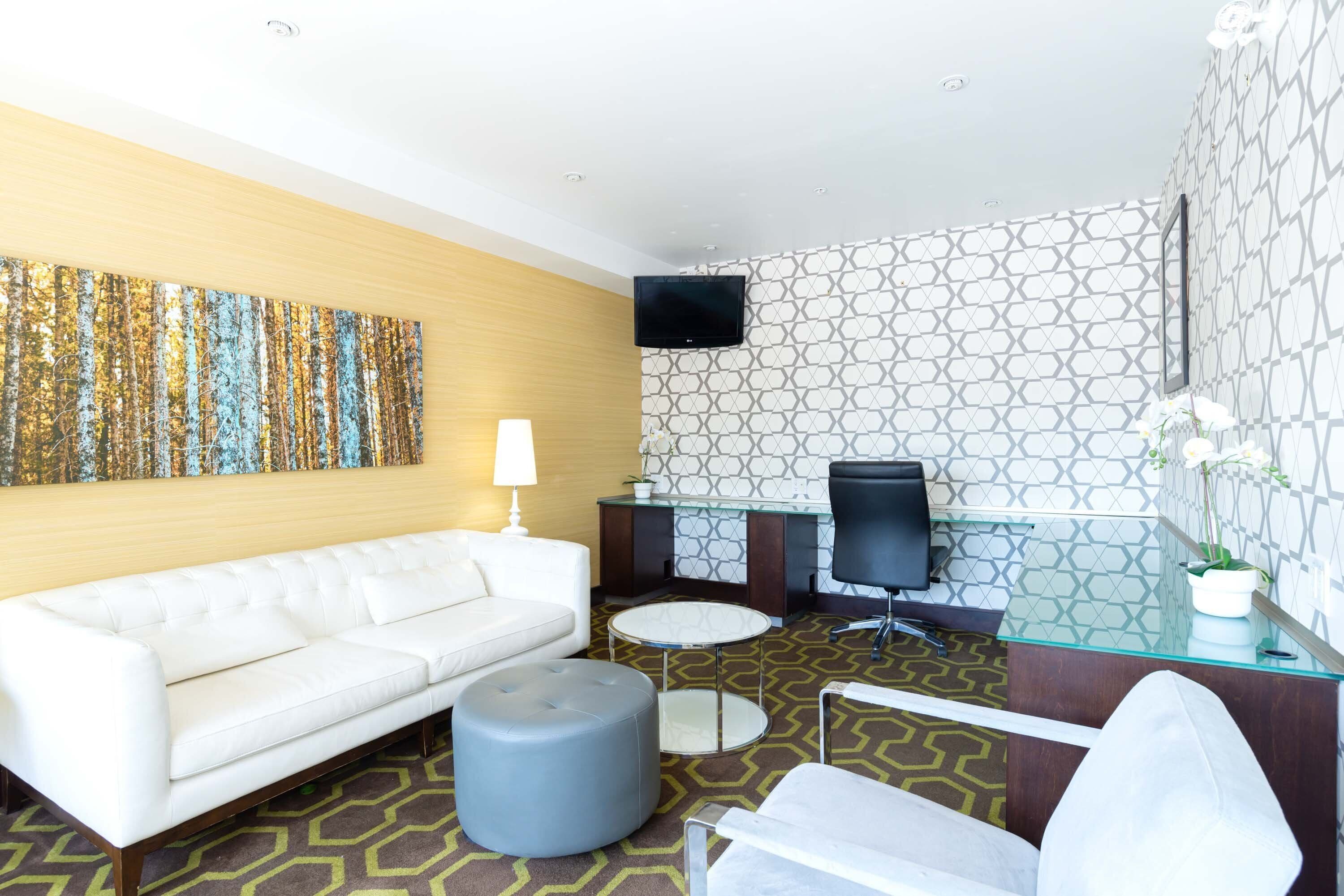 Ein modernes Hotelzimmer mit einem weißen Sofa, einem runden Hocker, einem Glastisch, einem Schreibtisch mit Stuhl, einem an der Wand montierten Flachbildfernseher und gemustertem Wandpapier.
