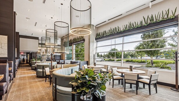 Un restaurante moderno con asientos tipo cabina y mesas, amplios ventanales que ofrecen vistas al exterior y una variedad de plantas que añaden verdor.