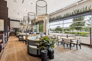 Un restaurante moderno con asientos tipo cabina y mesas, amplios ventanales que ofrecen vistas al exterior y una variedad de plantas que añaden verdor.