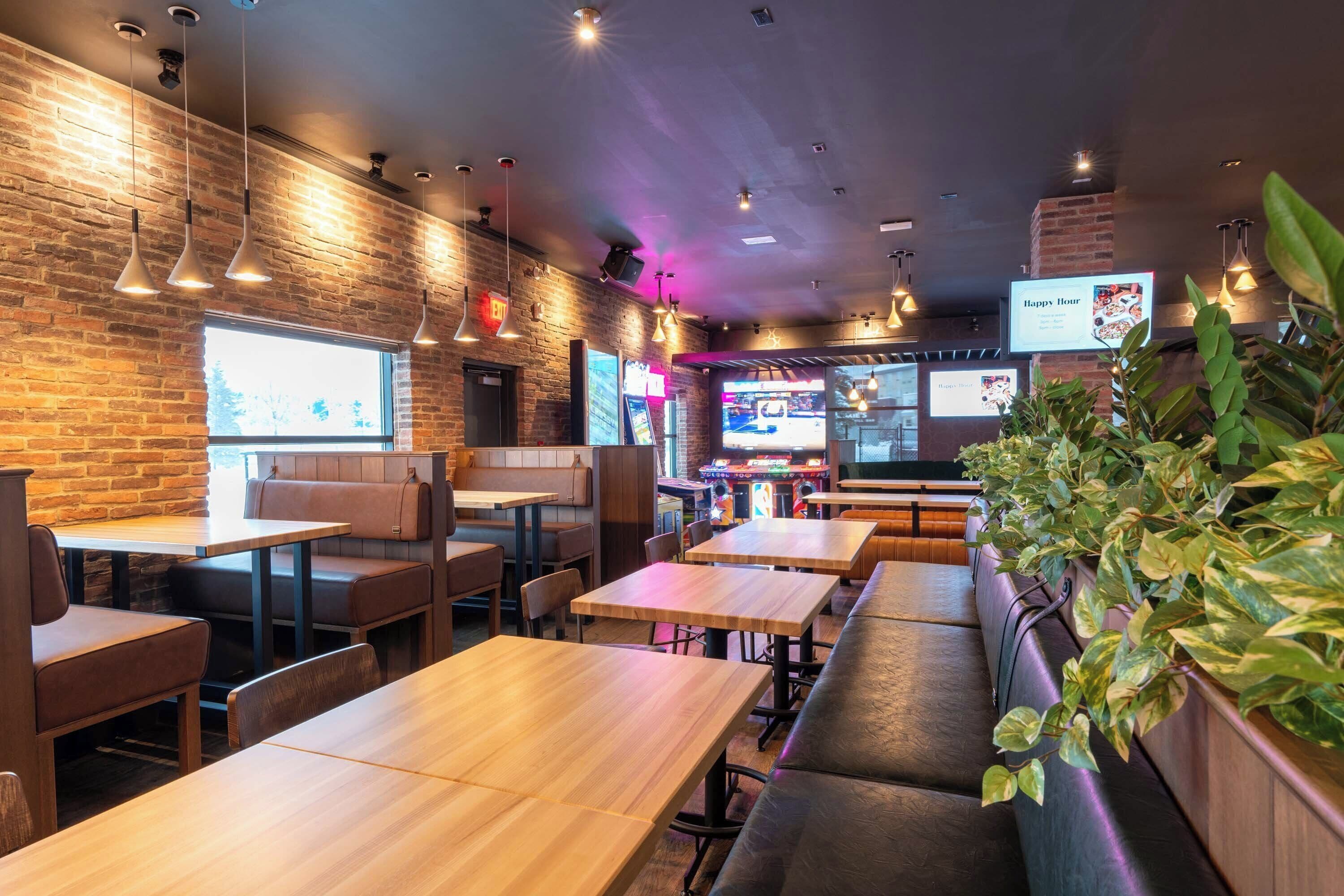 A restaurant interior with wooden tables and booths, brick walls, hanging pendant lights, and a large screen displaying a menu.