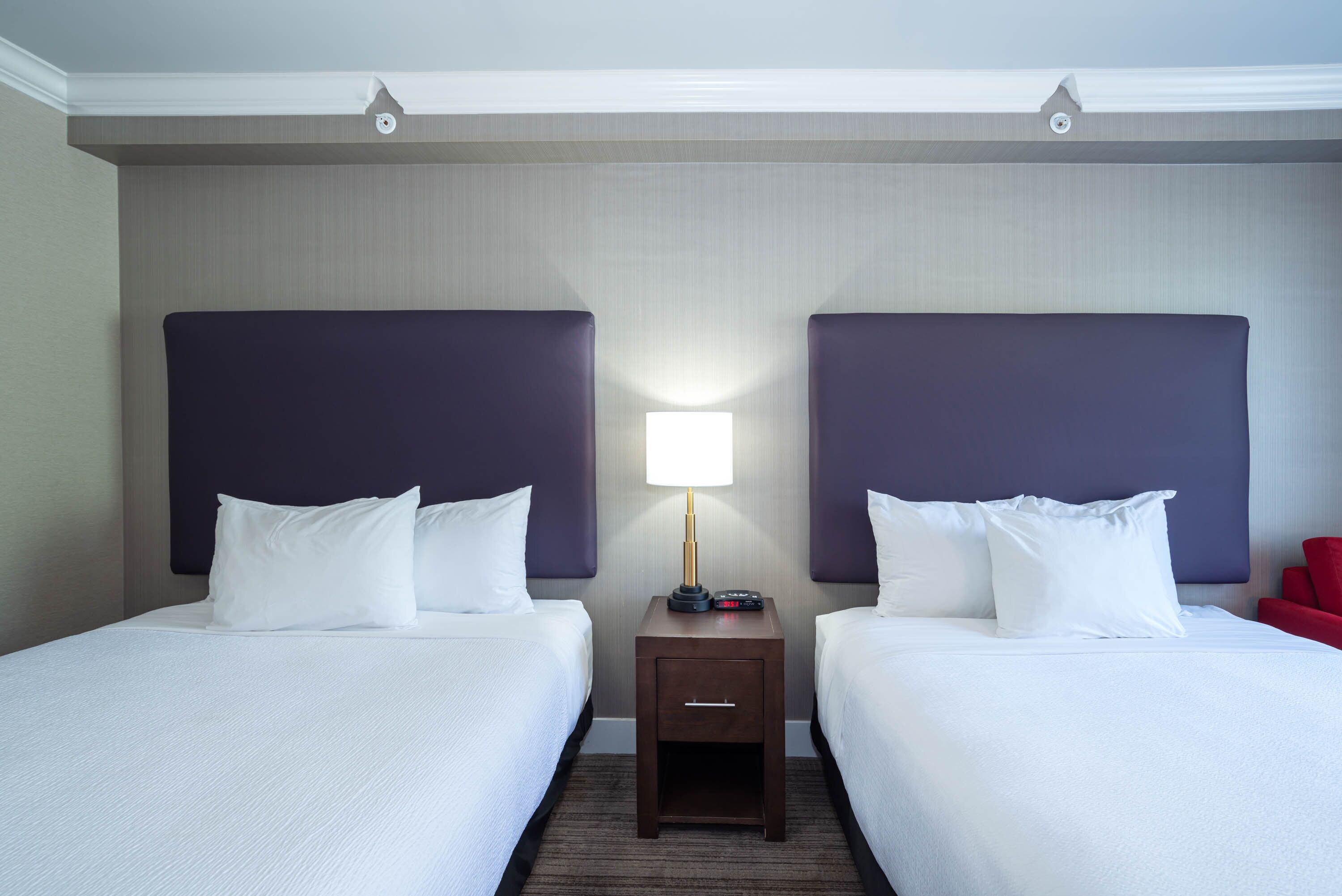 A hotel room with two beds, a nightstand with a lamp, and a red chair.