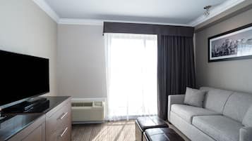 A hotel room with a flat-screen TV, a grey sofa, a brown ottoman, a large window with curtains, and a framed cityscape picture on the wall.