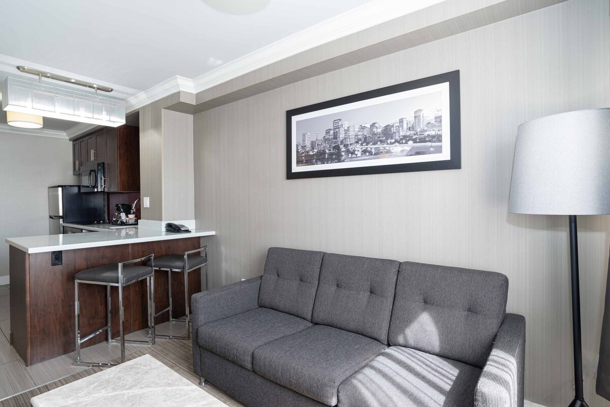 Una sala de estar moderna con un sofá gris, una pequeña cocina con un taburete de bar y un cuadro con una vista urbana enmarcada en la pared.
