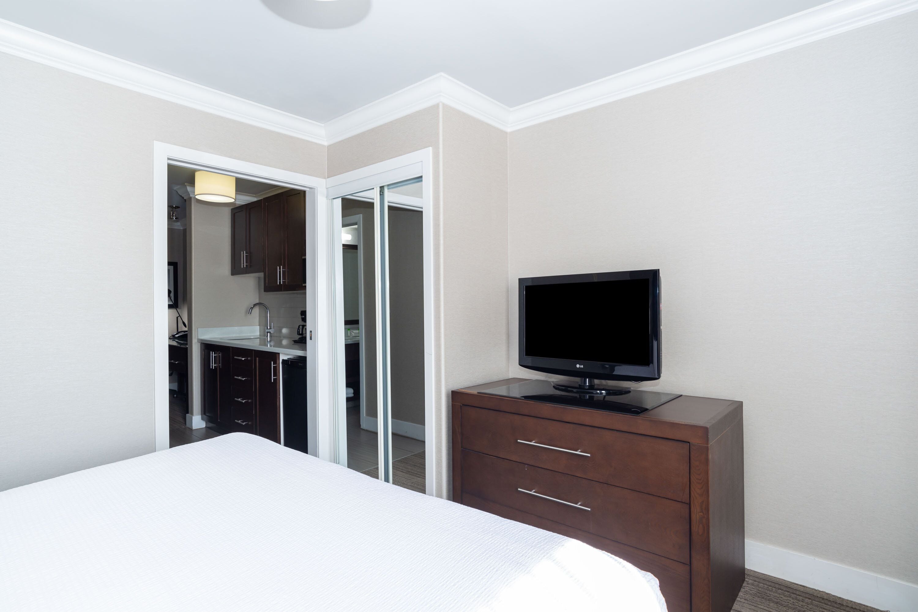 A hotel room with a bed, a TV on a wooden dresser, and a kitchenette in the background.