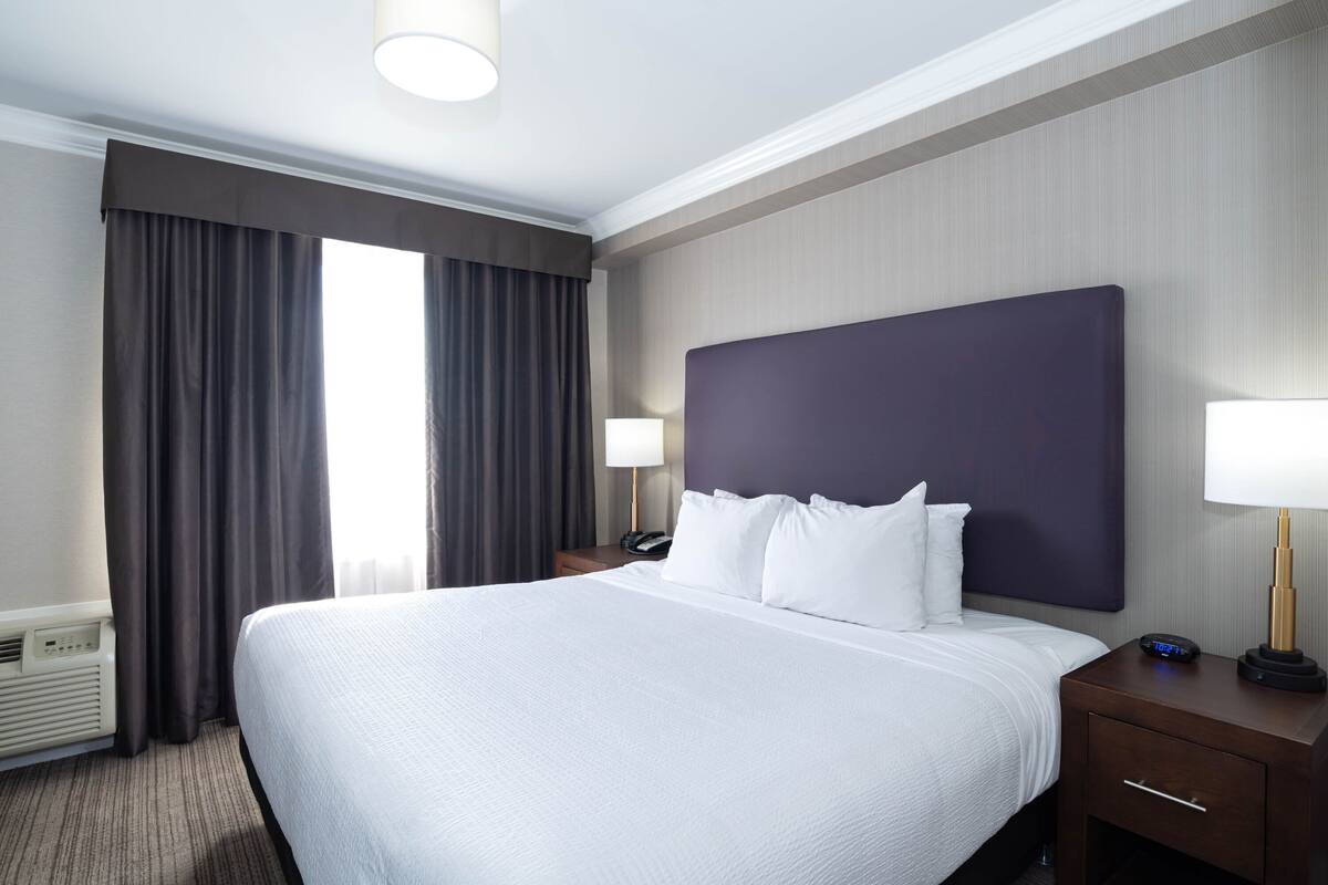 A hotel room with a large bed, two bedside tables with lamps, a heater, and a window with curtains.
