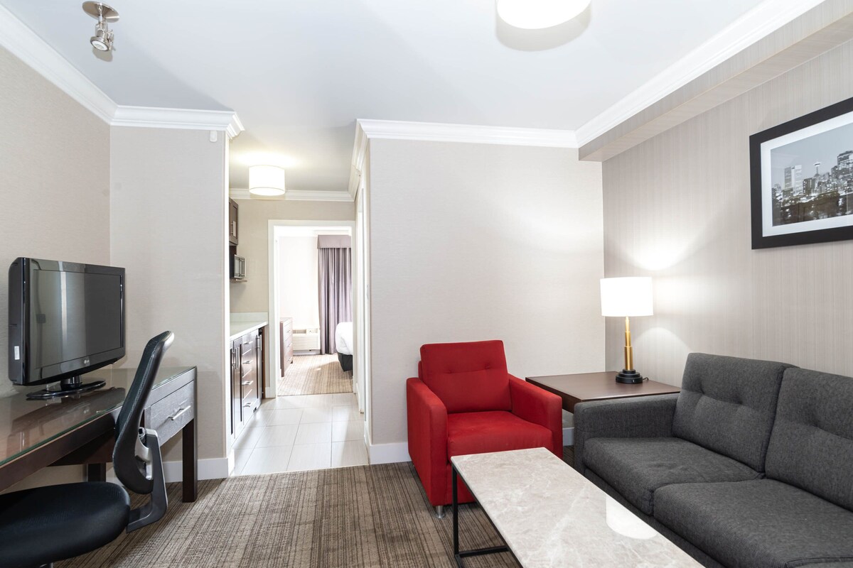 Habitación de hotel con televisor, escritorio, una silla roja, un sofá gris y una lámpara.