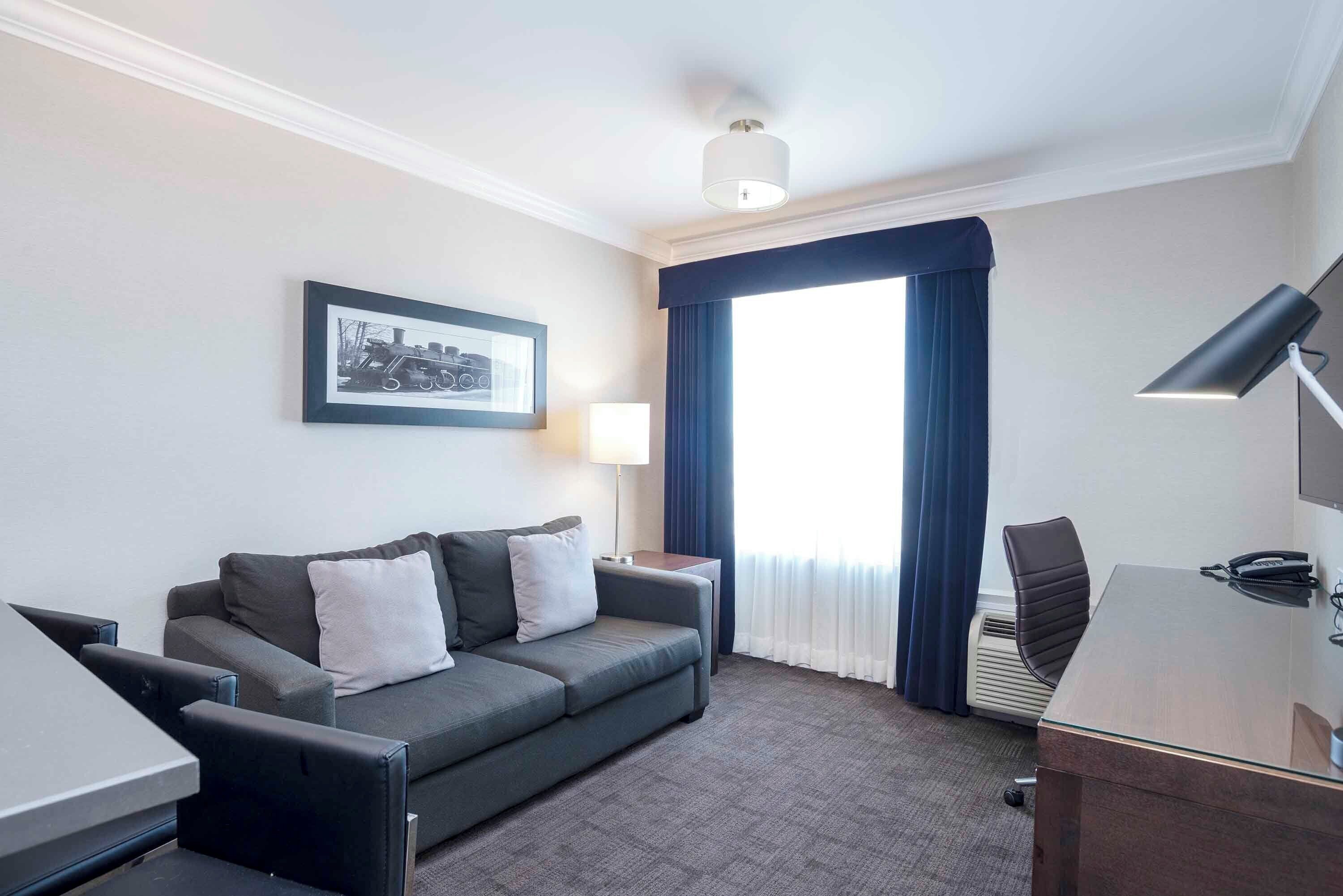 Habitación de hotel con sofá gris, escritorio, lámpara y una ventana grande con cortinas.