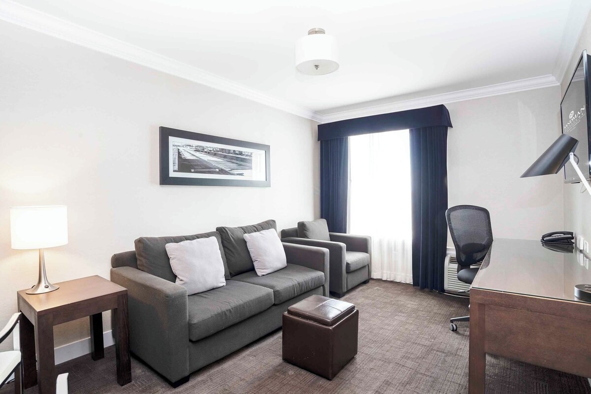 Een hotelkamer met een grijze bank, een klein tafeltje, een bureau en een stoel. Er hangt een lijstje op de muur en er is een raam met gordijnen.