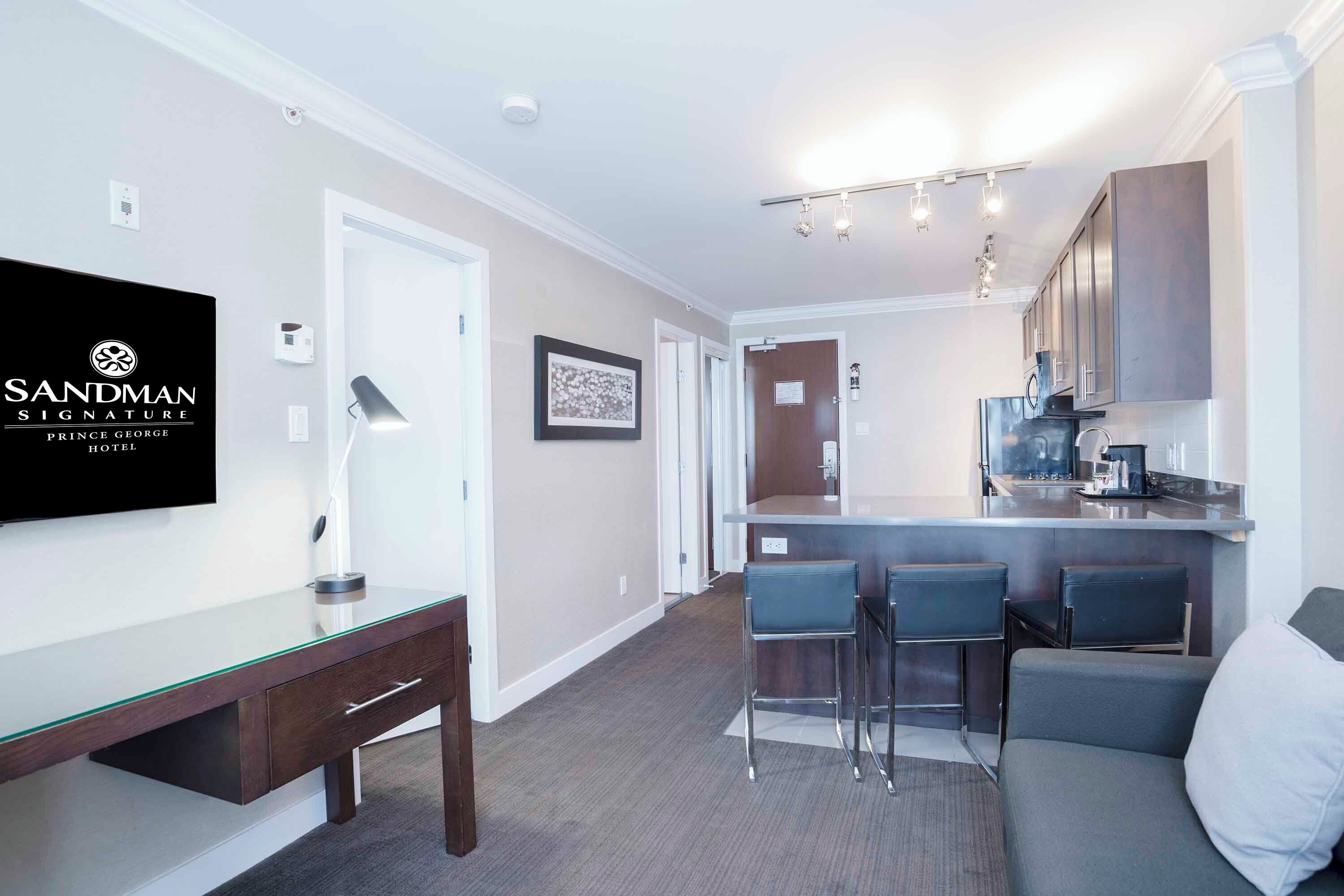 Una habitación de hotel moderna con un escritorio de vidrio, una zona de bar con taburetes, un sofá y una cocina con electrodomésticos de acero inoxidable.