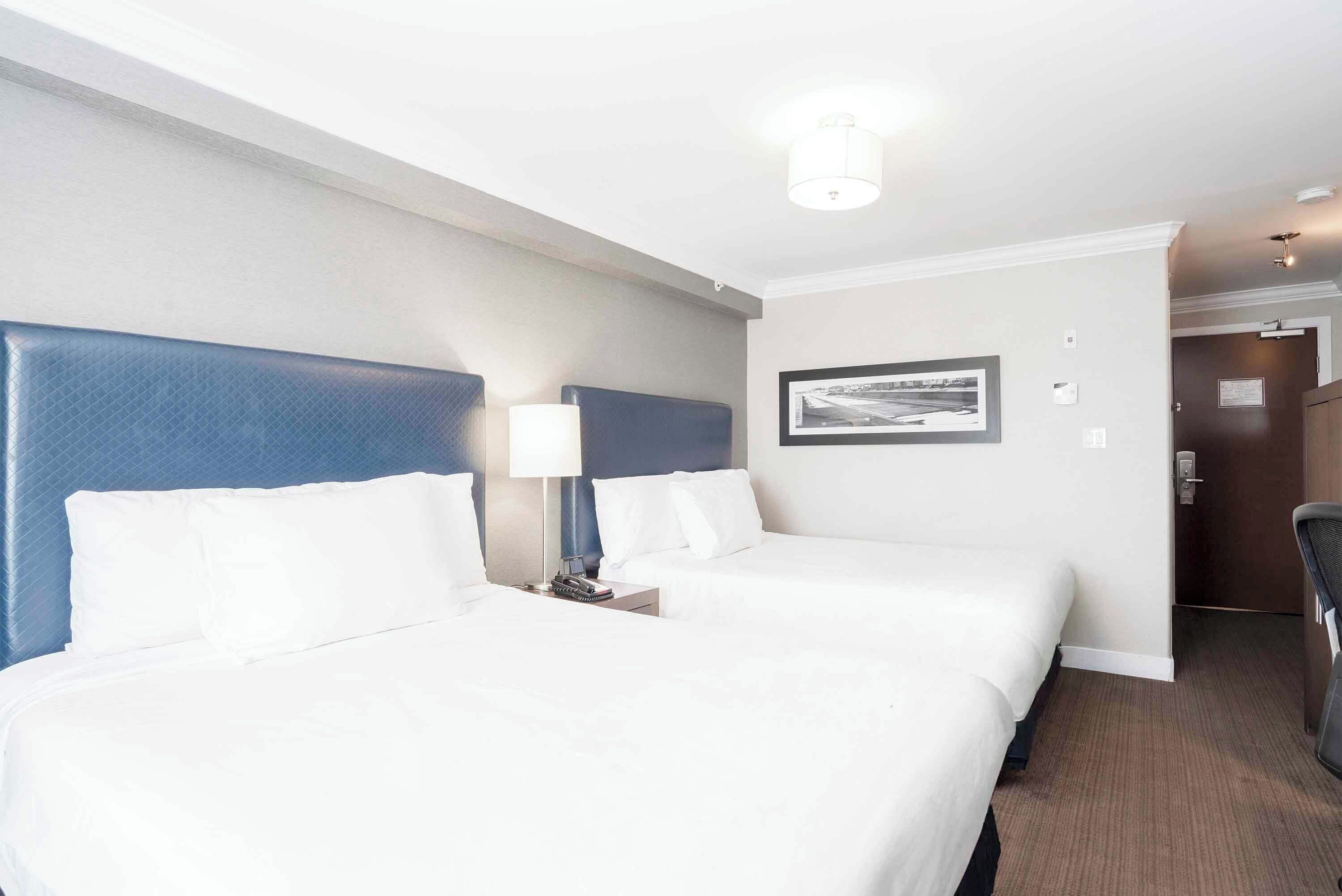 A hotel room with two beds, a headboard, a nightstand, a lamp, and a framed picture on the wall.