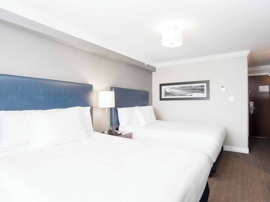 A hotel room with two beds, a headboard, a nightstand, a lamp, and a framed picture on the wall.