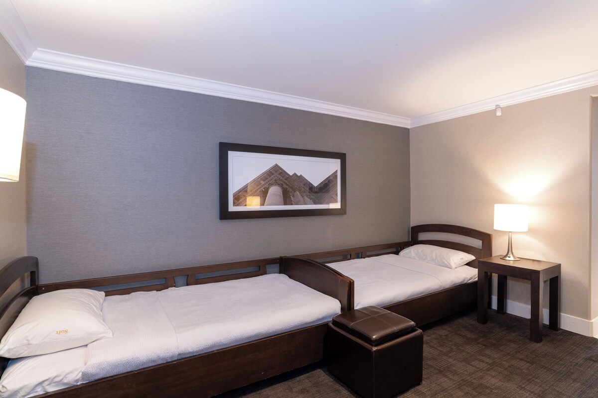 Een hotelkamer met twee enkele bedden, een tafeltje met een lamp, een lijst aan de muur en een vloerbedekking.