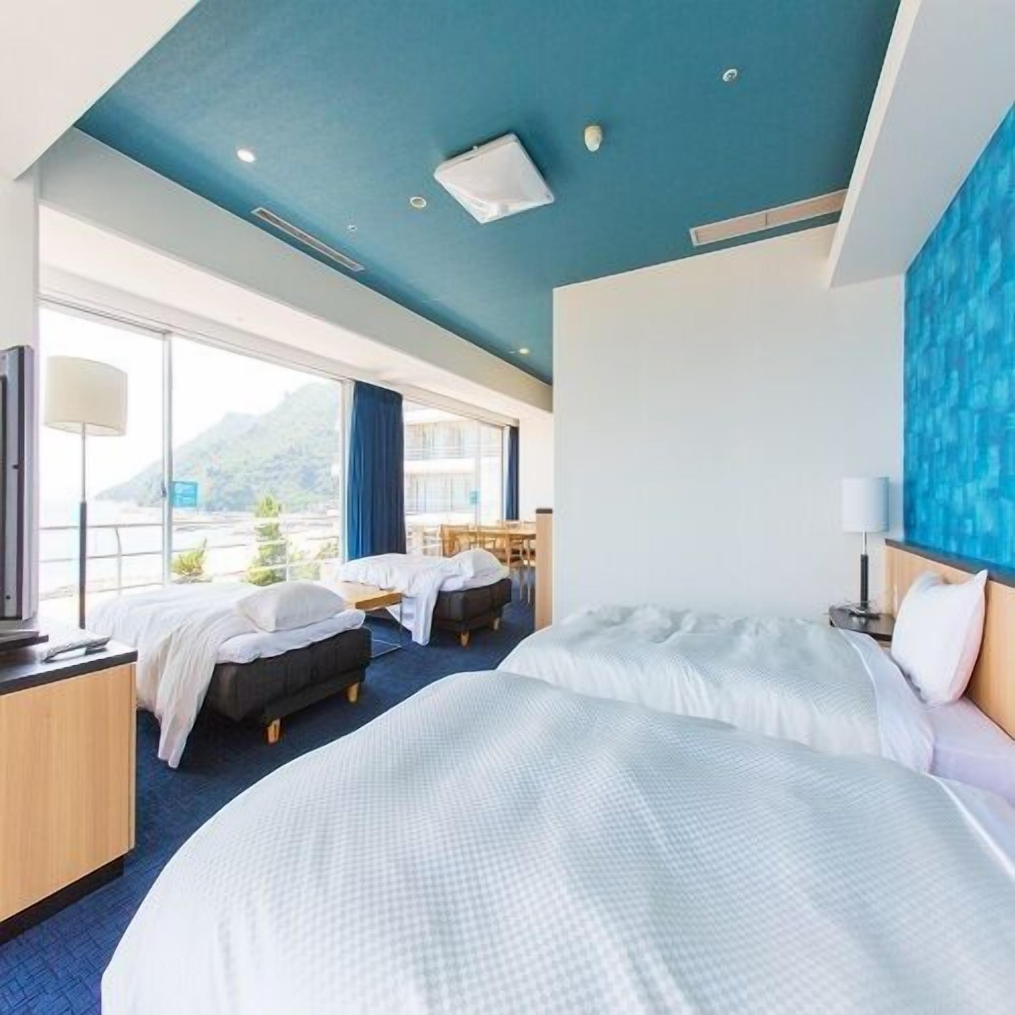 A hotel room with two beds, a desk, a TV, and a large window with a mountain view.
