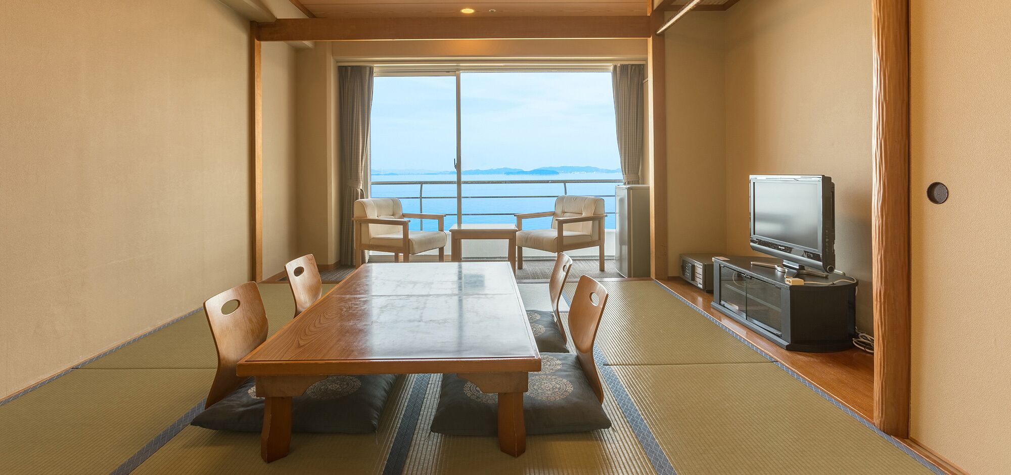 A room with a wooden table, chairs, a television, and a view of the sea.