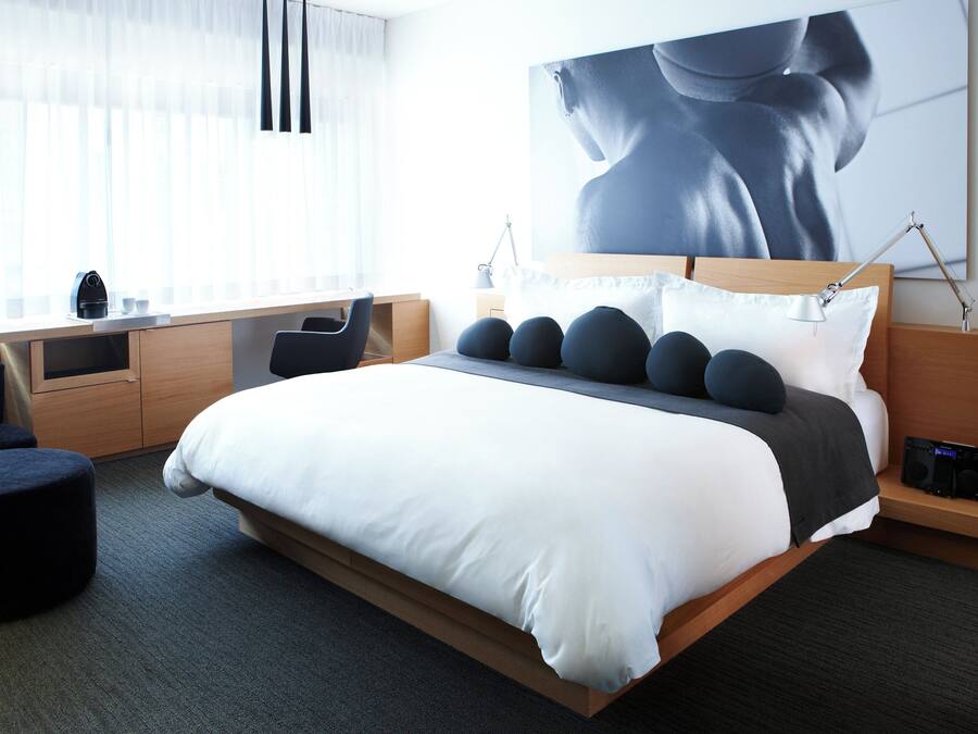 A modern hotel room with a large bed, a desk, and a chair. There is a large window with sheer curtains, a painting on the wall, and a bedside table with a lamp.
